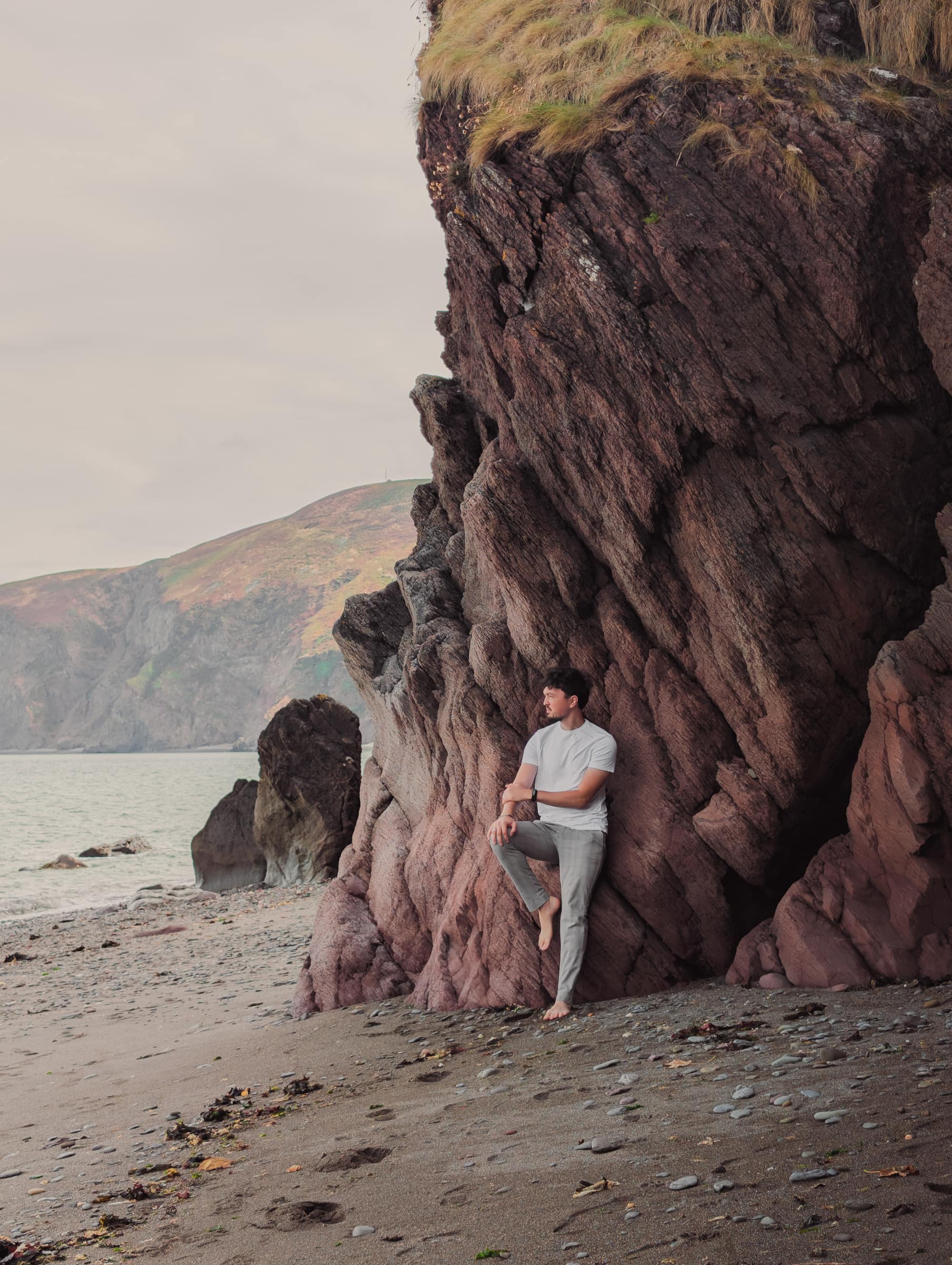Alex leaning against a huge rock formation on a quiet beach, sea and hills beyond.