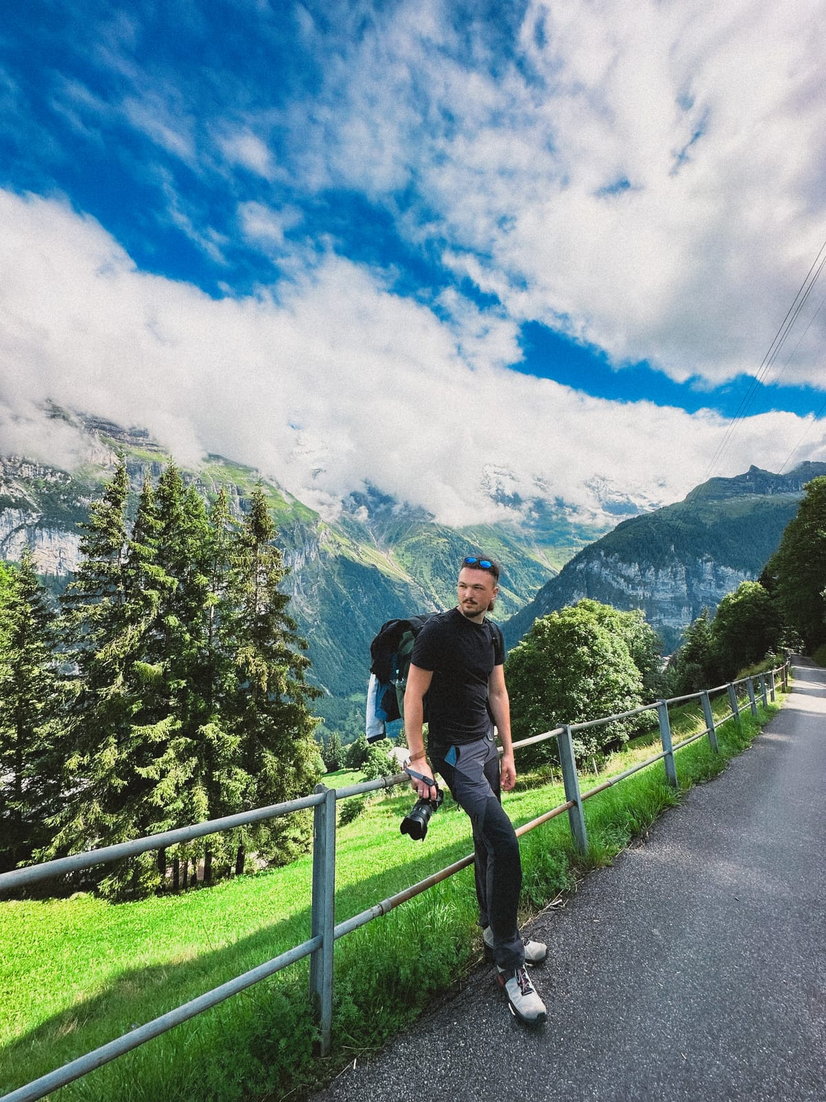 Alex on a sunny alpine road in Switzerland, black tee, camera in hand, green mountains and blue sky.