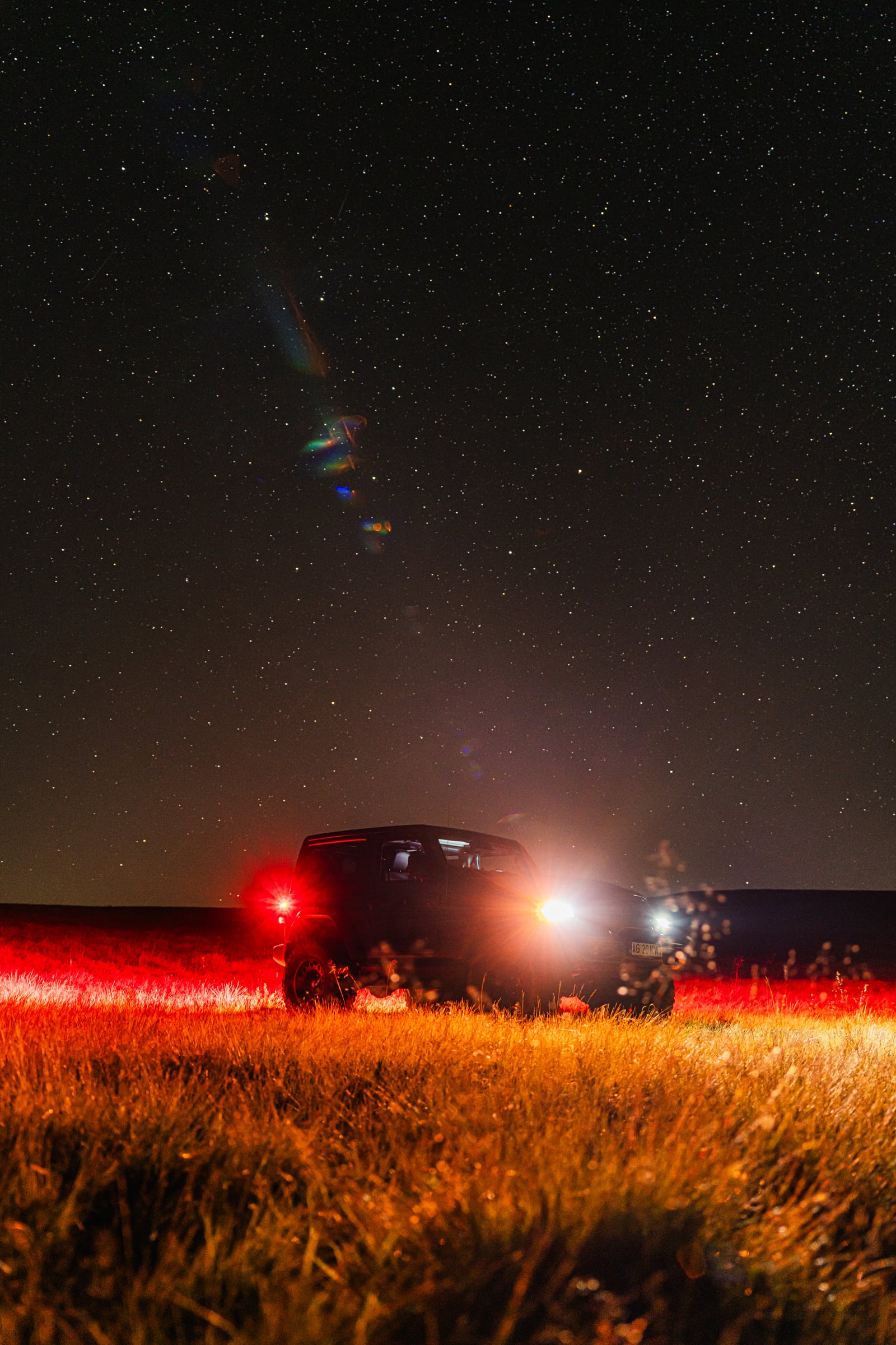 Night field under a starry sky, car parked in golden grass, red taillights glowing.