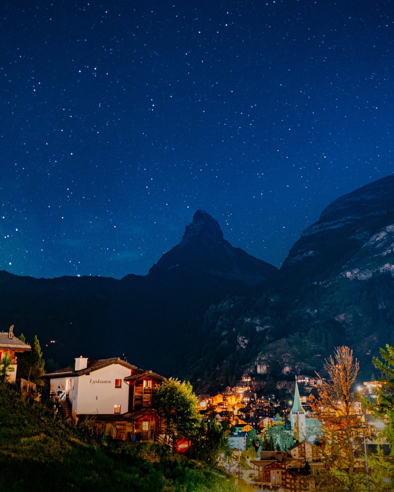 Night: village lights under a starry alpine peak.