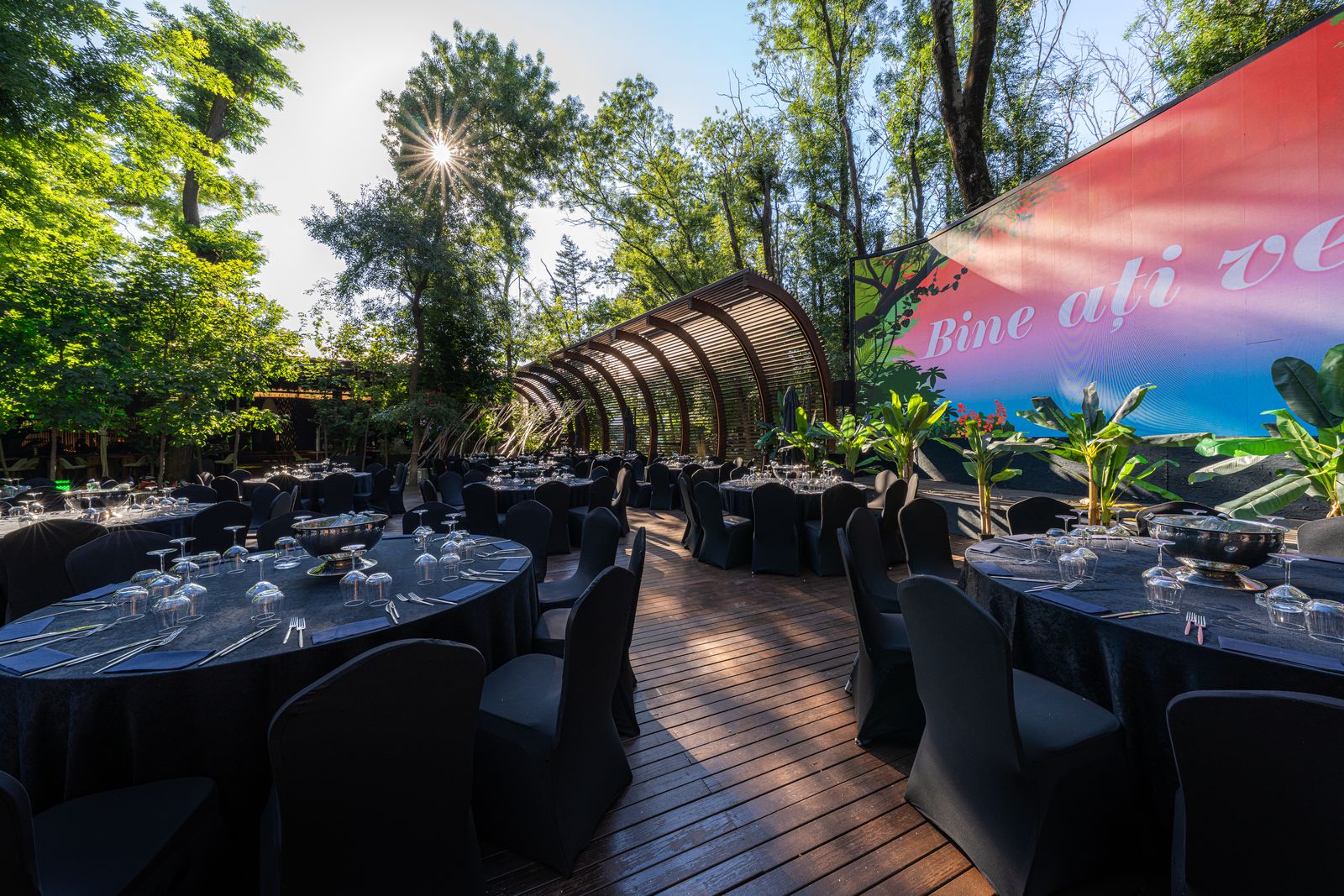Outdoor reception at the Face Convention Center — black-draped tables on a wooden deck under an arched pergola, "Bine ați venit" LED screen and sun flare through the trees.